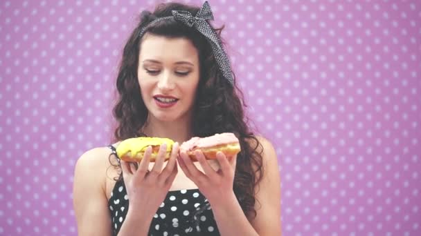 Heerlijk jong krullend meisje staan, kiezen tussen twee lekkere donuts in roze en gele glazuur in haar handen. - Video