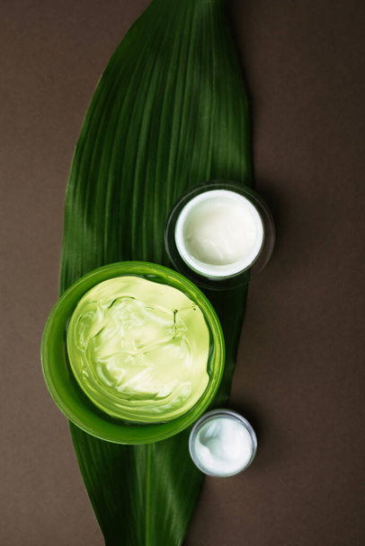 Jars of moisturizing white cream and green aloe gel on a palm leaf. Different moisturizers for different areas of the face on a brown background with copyspace. - Photo, Image