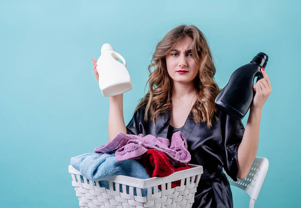 Conceito de lavandaria. dona de casa confusa segurando uma cesta de roupas e garrafas de detergente pronto para lavanderia isolada em fundo azul com espaço de cópia
 - Foto, Imagem