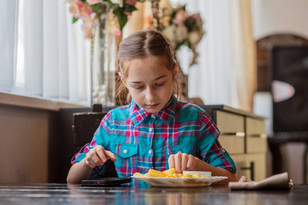 Een meisje dat frietjes eet. Meisje dat frietjes eet in een café. Een meisje van negen gaat lunchen met frietjes. Frietjes, saus, lunch, eten. Een schoolmeisje eet en kijkt naar een smartphone. - Foto, afbeelding