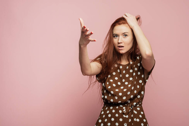 The red-haired girl is surprised and shocked. Woman experiences an emotion of rage on a pink background in the studio. - Foto, Imagen