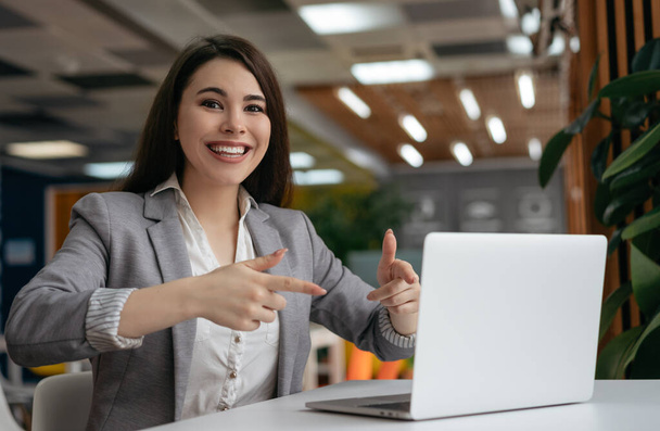 Happy emotional woman showing fingers on laptop computer, looking at camera and smiling. Online shopping, cash back, discount sales, hot deal concept - 写真・画像