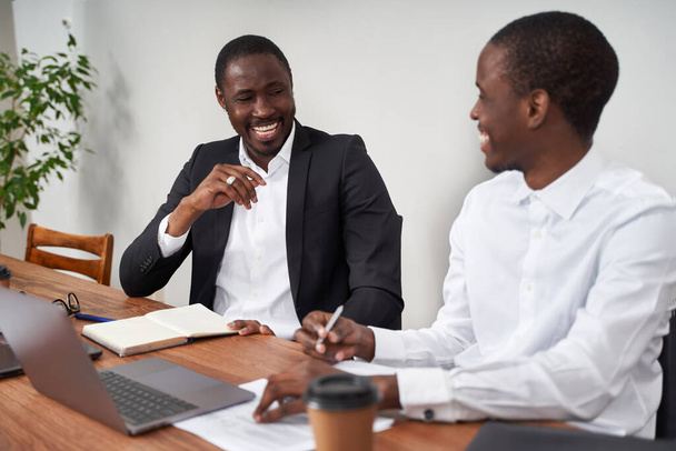 Śmiejący się afro-amerykańscy biznesmeni omawiający papierkową robotę ze współpracownikiem przy stole w biurze - Zdjęcie, obraz