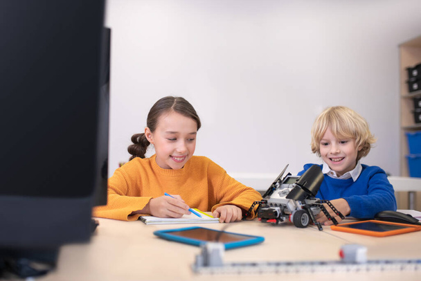 Alumnos sentados en el escritorio, tomando notas, jugando con el kit de construcción, sonriendo
 - Foto, Imagen