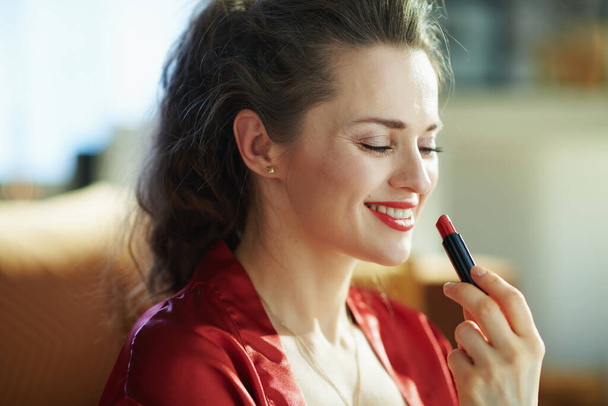 smiling modern housewife in black body lingerie and red bathrobe in the modern house in sunny day applying red lipstick. - Fotoğraf, Görsel