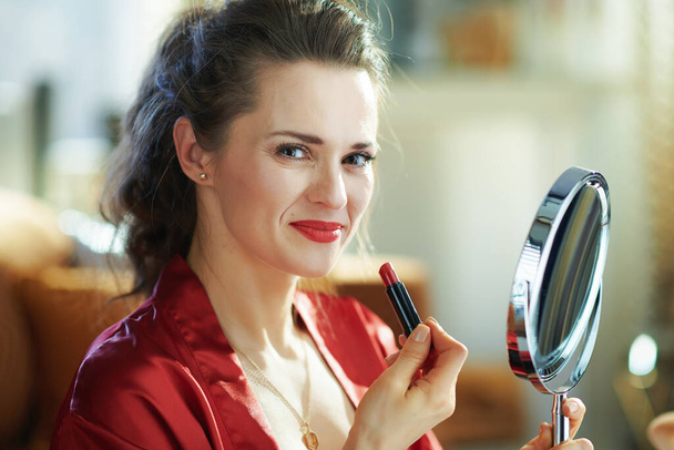 Portrait of smiling trendy middle age woman in black body lingerie and red bathrobe with red lipstick and mirror in the modern house in sunny day. - Foto, afbeelding