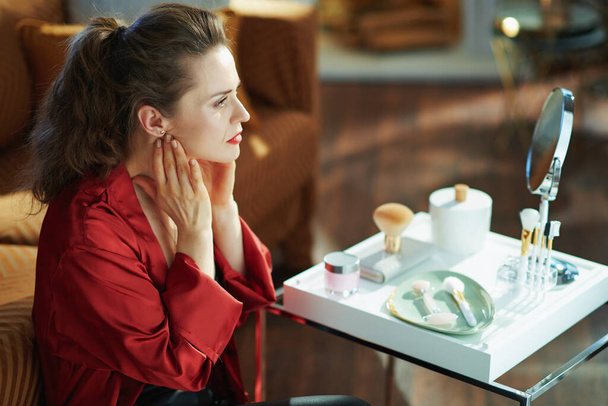 concerned trendy middle age housewife in black body lingerie and red bathrobe looking in mirror and checking wrinkles near table with toiletries in the modern house in sunny day. - Fotoğraf, Görsel