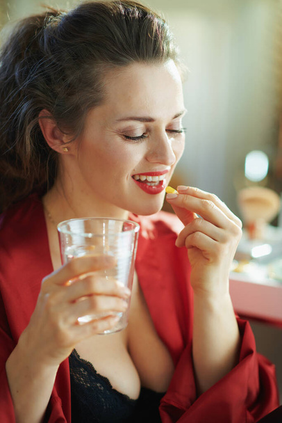 glückliche elegante Frau in schwarzer Körperunterwäsche und rotem Bademantel mit einer Tasse Wasser, die an sonnigen Tagen im modernen Wohnzimmer Tabletten einnimmt. - Foto, Bild