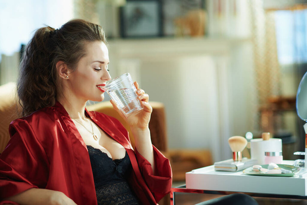 happy middle age woman in black body lingerie and red bathrobe drinking cup of water near table with toiletries in the modern house in sunny day. - 写真・画像