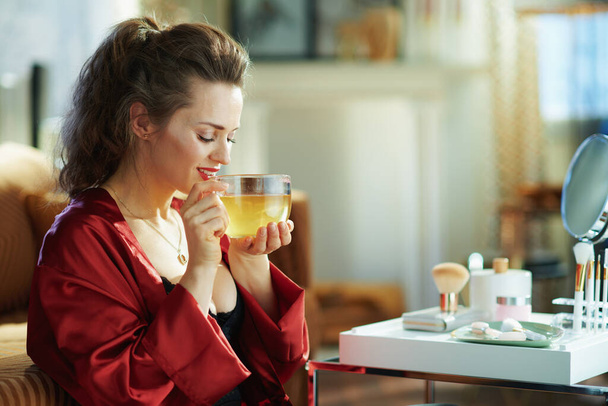relaxed elegant woman in black body lingerie and red bathrobe with cup of green tea near table with toiletries in the modern living room in sunny day. - Фото, зображення