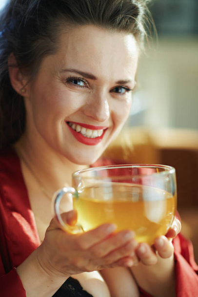 Portrait de heureuse femme d'âge moyen en lingerie corps noir et peignoir rouge avec tasse de thé vert dans le salon moderne dans la journée ensoleillée
. - Photo, image