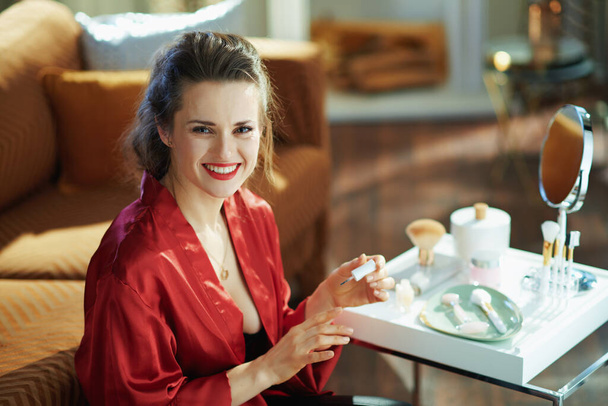 happy elegant middle age housewife in black body lingerie and red bathrobe applying nail polish near table with toiletries in the modern living room in sunny day. - Foto, imagen