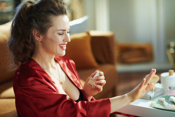 happy trendy middle age housewife in black body lingerie and red bathrobe with nail polish bottle looking at nails at modern home in sunny day. - Фото, зображення