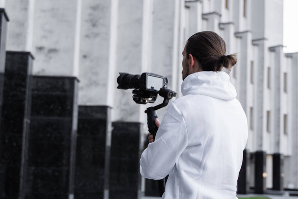 Young Professional videographer holding professional camera on 3-axis gimbal stabilizer. Pro equipment helps to make high quality video without shaking. Cameraman wearing white hoodie making a videos. - Zdjęcie, obraz