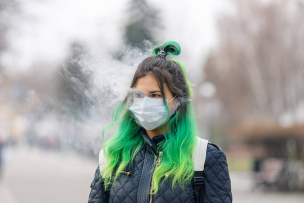Dziewczyna z zielonymi włosami w ochronnej masce medycznej i pali vape. - Zdjęcie, obraz