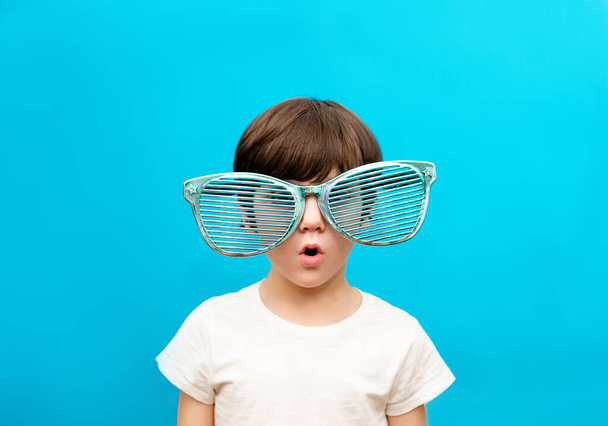 Веселый мальчик в больших очках выражает удивленное лицо, изолированное на синем фоне - Фото, изображение