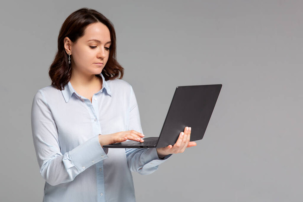 Charmante junge Frau, die freiberuflich arbeitet, fernab vom Büro, Laptop in der Hand, Aufsatz an der Universität schreiben, lächelnd nach links abbiegen, fröhlich grauen Hintergrund stehen, mit dem Computer im Internet surfen - Foto, Bild