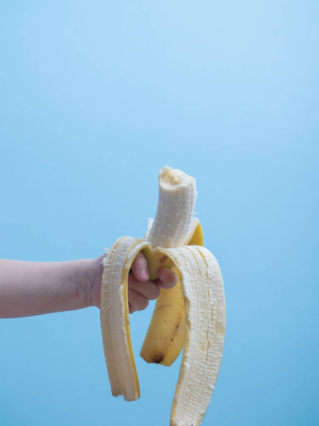 a childs hand holds a bitten banana on a blue backgroun - Фото, изображение