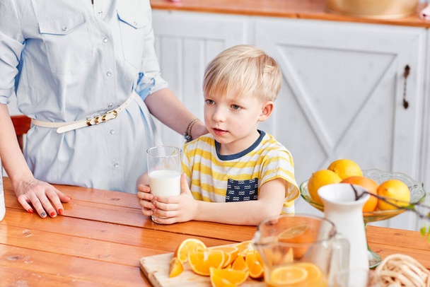Ten kluk pije mléko ze sklenice. Matka a syn se usmívají, když snídají v kuchyni. Máma nalévá mléko do skla - Fotografie, Obrázek