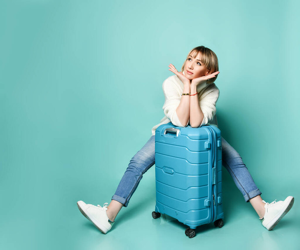 Blondes Mädchen in weißem Pullover, Jeans und Turnschuhen. Sitzt auf ihrem Koffer, stützt ihr Gesicht ab, posiert auf blauem Hintergrund - Foto, Bild