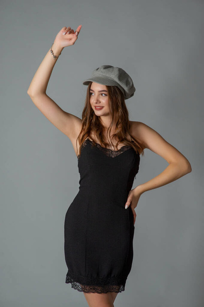 Glamorous young woman wears fashionable dress posing with fluttering hair at grey wall. Girl put forward hand in a black dress and a gray cap. Beautiful young girl with brown curly hairs. portrait - Valokuva, kuva