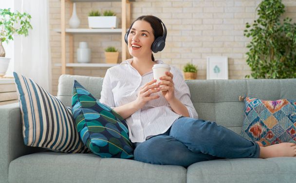 Cheerful young woman listening to music in headphones sitting on the sofa in the room at home. - Foto, Imagem
