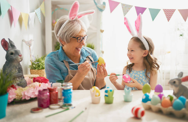Happy easter! A grandmother and her granddaughter painting Easter eggs. Happy family preparing for Easter. Cute little child girl wearing bunny ears on Easter day. - Photo, Image