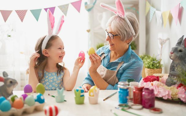 Happy easter! A grandmother and her granddaughter painting Easter eggs. Happy family preparing for Easter. Cute little child girl wearing bunny ears on Easter day.                                - Foto, imagen