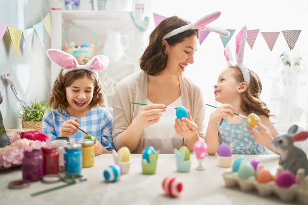 Happy holiday! A mother and her daughters are painting eggs. Family preparing for Easter. Cute little children girls are wearing bunny ears.                                 - Photo, Image
