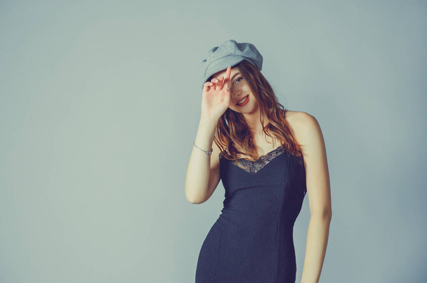 Glamorous young woman wears fashionable dress posing with fluttering hair at grey wall. Girl put forward hand in a black dress and a gray cap. Beautiful young girl with brown curly hairs. portrait - Photo, Image