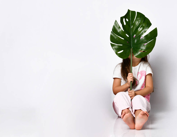 little girl in summer pastel clothes sits on the floor, covering her face with a large crested sheet of tropical raster - Foto, imagen