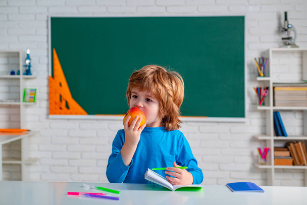 Elementary school and education. Happy school kid eat apple. Portrait of Pupil hold apple in classroom. Chalkboard copy space. - 写真・画像