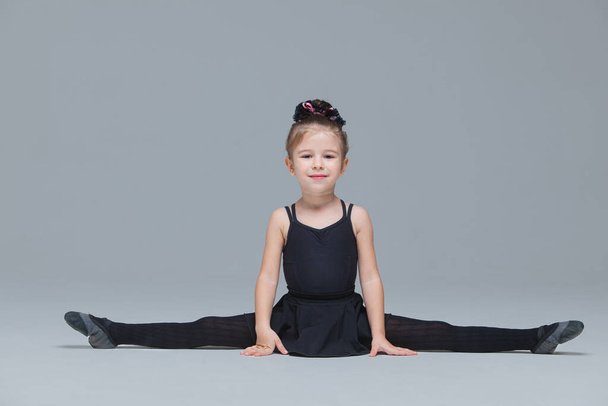 Beautiful little gymnast in black sportswear shows exercises on grey background. Flexible girl doing the splits to become ballet dancer - Foto, afbeelding