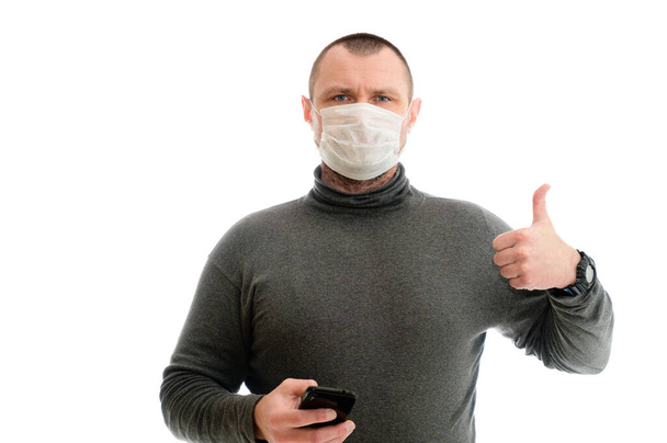 Concept isolate Caucasian man in a medical mask in a gray jacket on a white background with a smartphone shows ok with hands - Foto, imagen