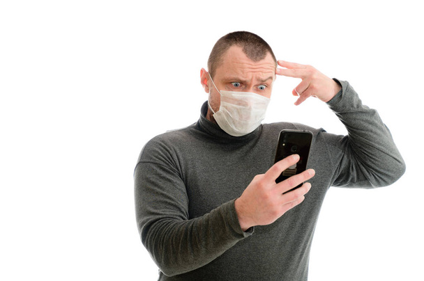 The model stands right in front of the camera. With smartphone shows emotions with hands concept isolate Caucasian man in a medical mask in a gray sweater on a white background - Zdjęcie, obraz