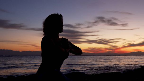 A silhueta da menina senta-se na pose namaste que medita pelo mar no por do sol
. - Foto, Imagem