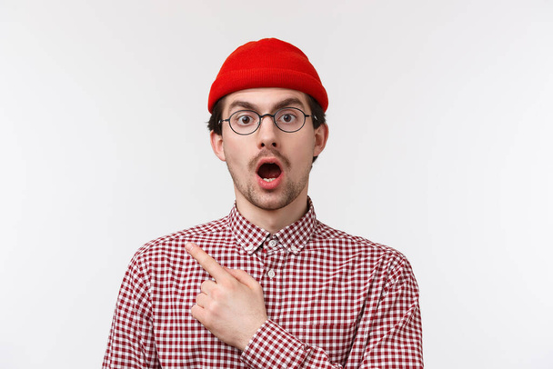Překvapený a ohromený pohledný vousatý hipster muž v brýlích a červené čepici, s otevřenými ústy a klesající čelistí udýchaný, uviděl něco šokujícího, ukazující levý horní roh, bílé pozadí - Fotografie, Obrázek