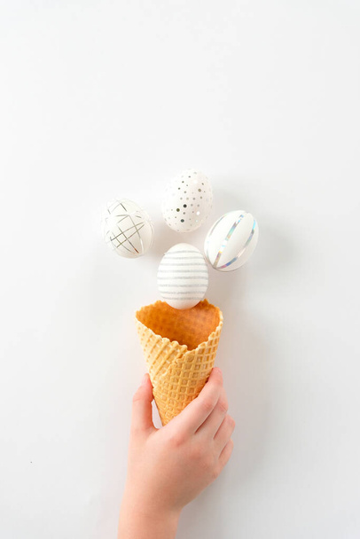 Hand holds a waffle cone with white paper flowers and white easter eggs with silver decor on white background. Happy Easter concept. Long format for web - Фото, изображение