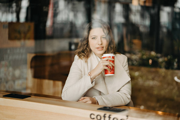 Chica mira a través de la ventana de una cafetería y sostiene el café en sus manos
 - Foto, imagen