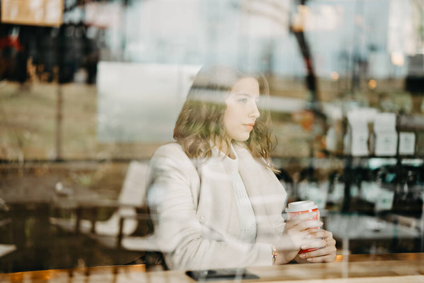 Chica mira a través de la ventana de una cafetería y sostiene el café en sus manos
 - Foto, Imagen