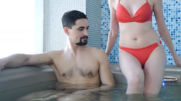 Wellness spa centre in luxury hotel. Couple relaxing in jacuzzi. Attractive slim woman in red bikini swimsuit entering the water in pool. 4 k footage - Video, Çekim