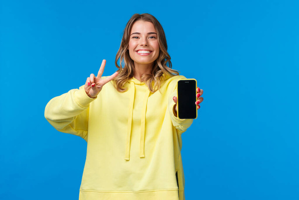 Carino sorridente ragazza bionda felice in felpa gialla con cappuccio che mostra il suo blog online o pagina di social media sul display del telefono cellulare, fare gesto di pace e sorridente, stand sfondo blu
 - Foto, immagini