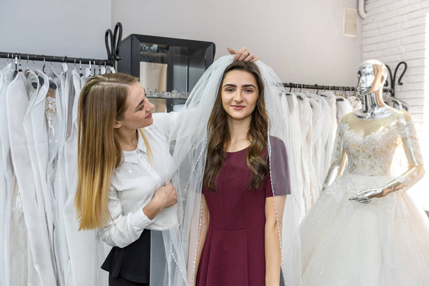 Saleswoman fitting veil for young bride in store - Valokuva, kuva