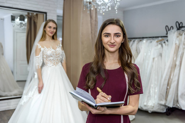Дизайнер с блокнотом и невестой в свадебном платье позади
 - Фото, изображение