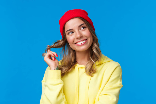 Close-up portrait dreamy silly cute european female in red hipster beanie, yellow hoodie, rolling curl on hair and gazing upper right corner thoughtful, standing blue background - Foto, afbeelding