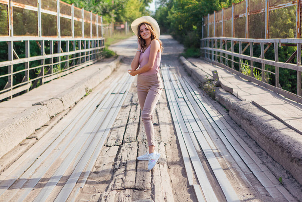 Warm summer portrait of a young beautiful woman wearing cowboy hat posing on the old rusty bridge during sunset. - Foto, Imagem