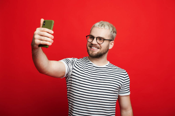 Blonder Kaukasier, der durch eine Brille blickt, macht mit seinem Handy ein Selbstporträt, während er auf rotem Hintergrund posiert - Foto, Bild