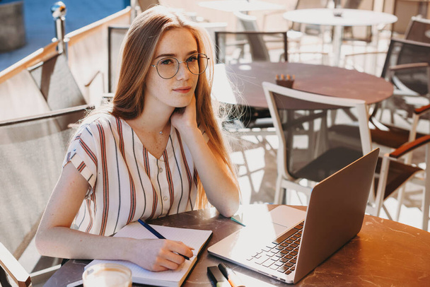 Kaukasische Frau mit roten Haaren und Sommersprossen bei einer Kaffeepause in einem Café, während sie einen Laptop benutzt und ein paar Notizen macht - Foto, Bild