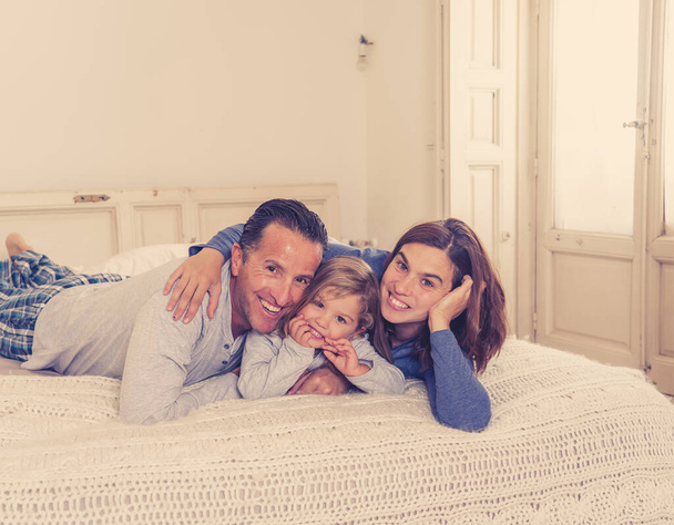 Lifestyle portrait of beautiful happy family relaxing and spending time together in bed at home. Mother, father, and cute toddler daughter having fun kissing hugging and bonding in the morning. - Photo, Image