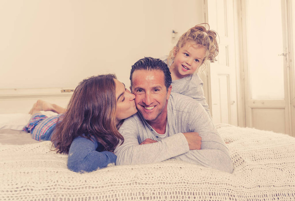Portret życia pięknej szczęśliwej rodziny relaksujący i spędzający czas razem w łóżku w domu. Mama, ojciec, i słodkie dziecko córka mając zabawy całowanie przytulanie i więzi rano. - Zdjęcie, obraz